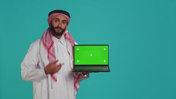 Muslim adult shows greenscreen laptop with blank mockup layout on display, copyspace template. Arab guy wearing gown and headscarf, presenting isolated chromakey screen on pc. video