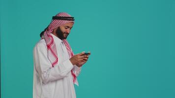 gelukkig persoon hebben pret met spel Aan zijn smartphone apparaat, vervelend traditioneel Arabisch kleding. in de studio, moslim model- genieten van e sport toernooi met mobiel telefoon videospellen. video