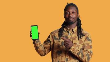 Smiling man points to smartphone with greenscreen on camera, showing blank mockup template on mobile device display. Young adult presenting isolated chromakey layout on phone screen. video