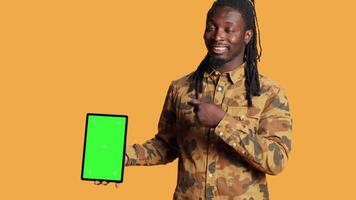 African american man presents greenscreen on tablet, showing isolated copyspace screen and smiling in front of camera. Young person holding mobile gadget with blank mockup display layout. video