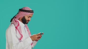 Arabic person playing mobile videogames on smartphone with tensed expression, wears islamic headscarf and thobe. Muslim gamer enjoying online gaming experience on phone app in studio. video