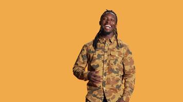 Cheerful adult enjoying shoot and laughing on camera, making jokes in studio and acting playful. Silly positive african american guy having fun over orange background, relaxed casual person. video