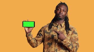 Cheerful adult holds smartphone with greenscreen in studio, presenting isolated copyspace display on mobile phone layout. Young person showing blank chromakey mockup on device. video