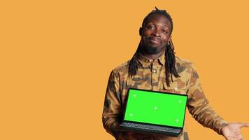 Young guy showing pc with greenscreen layout on camera, pointing at isolated chromakey display. African american man presenting laptop with blank copyspace mockup screen in studio. video
