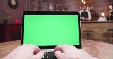 Mano pov Disparo de masculino manos mecanografía en un ordenador portátil con verde pantalla. en el antecedentes un barman es trabajando a el bar mostrador video