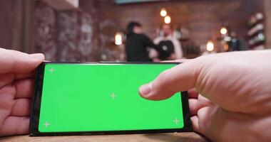 hombre deslizar en un verde pantalla teléfono en un ocupado restaurante, café tienda o pub. un barman es hablando con un cliente en el antecedentes video