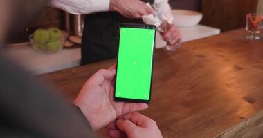 homme balayages sur une téléphone avec vert écran tandis que séance à le comptoir et le barman est nettoyage une verre dans le Contexte video