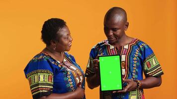 Married people pointing at tablet with greenscreen layout, looking at isolated mockup template on gadget. Ethnic couple showing modern blank copyspace screen on mobile device. video