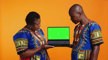 Ethnic couple showcasing laptop with greenscreen display, presenting isolated copyspace template and posing on camera. African american man and woman pointing at blank mockup layout. video