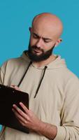 Vertical Video Middle eastern adult creating a checklist on clipboard papers, being focused on trying to plan tasks on an organizer in studio. Modern person looking at files and listing things to do. Camera 1.