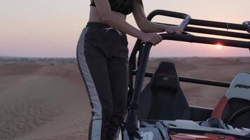 Young woman in sportswear standing on a buggy in desert sand against sunset background video