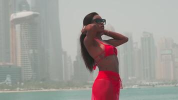 Dubai, UAE - 5 22 2021. A young swarthy woman in a bikini and a light red dress walks barefoot on the rocks against the backdrop of the sea and skyscrapers of Dubai. video