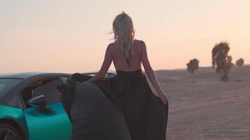 une Jeune femme avec blond cheveux flottant dans le vent et une longue noir robe des stands pieds nus sur le le sable suivant à un coûteux des sports voiture video