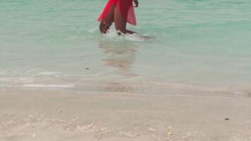 een jong donker vrouw in een bikini en een licht rood jurk wandelingen blootsvoets Aan een zanderig strand in de surfen water video