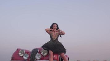 A young spectacular woman in a black dress and high-heeled shoes stands on the road next to a red vintage car in the middle of the desert video