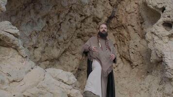 A young bearded man with a staff in his hands in the clothes of a shaman prays on a rock among the sandy dunes of the desert video