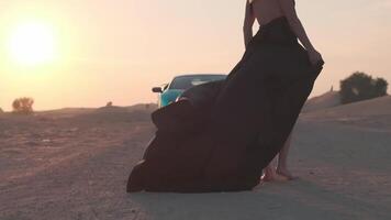 een jong vrouw met blond haar- fladderend in de wind en een lang zwart jurk staat blootsvoets Aan de zand De volgende naar een duur sport- auto video