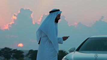 A young bearded businessman in national Arab clothes is talking on the phone next to an expensive premium car. video