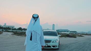 een jong gebaard zakenman in nationaal Arabisch kleren staat De volgende naar een wit premie auto. video