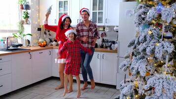 gelukkig traditioneel familie van pa, mama, dochter in feestelijk de kerstman hoeden grappig en pret dansen in een wit keuken met een Kerstmis boom en decor. nieuw jaar, familie waarden. video