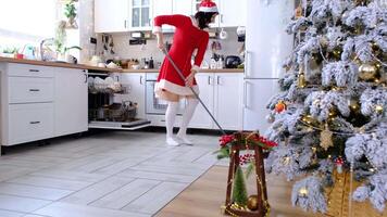 A woman in a red dress and Santa hat cleans the floor with a mop in a white kitchen with Christmas decor and a Christmas tree. Cleaning the house and preparing for the holidays Christmas and New Year video