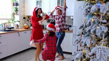 gelukkig traditioneel familie van pa, mama, dochter in feestelijk de kerstman hoeden grappig en pret dansen in een wit keuken met een Kerstmis boom en decor. nieuw jaar, familie waarden. video