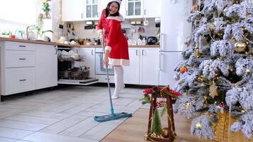 A woman in a red dress and Santa hat cleans the floor with a mop in a white kitchen with Christmas decor and a Christmas tree. Cleaning the house and preparing for the holidays Christmas and New Year video