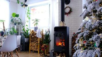 Metall black stove, burning hearth fireplace in white Festive interior of house is decorated for Christmas and New Year, Christmas tree. firewood in the woodpile, cozy and heating of home video
