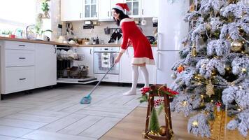 en kvinna i en röd klänning och santa hatt rengör de golv med en mopp i en vit kök med jul dekor och en jul träd. rengöring de hus och framställning för de högtider jul och ny år video