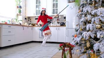 un mujer en un rojo vestir y Papa Noel sombrero limpia el piso con un fregona en un blanco cocina con Navidad decoración y teniendo divertido, baile, gracioso jugando me gusta un guitarra, canto dentro un palo me gusta un micrófono video