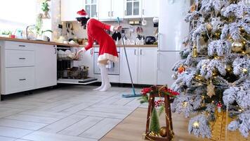 Woman in red dress and Santa hat cleans the floor with mop and closes dishwasher in white kitchen with Christmas decor and having fun, dancing, singing into a stick like a microphone video