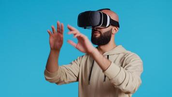 Middle eastern adult using virtual reality glasses in studio, having fun with metaverse artificial intelligence on vr headset. Young person posing over blue background, modern goggles. Camera 2. video