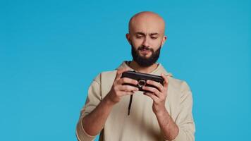 alegre musulmán hombre jugando móvil vídeo juegos en estudio, disfrutando en línea competencia utilizando teléfono inteligente aplicación Arábica jugador teniendo divertido con juego de rol desafío concurso. cámara 2. Mano disparo. video