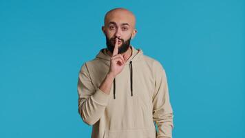 Middle eastern man doing hush silence symbol in studio, asking to keep a secret over blue background. Arab person creating mute gesture to be quiet in front of camera. Camera 1. video