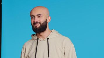 Middle eastern guy greeting people in front of camera, waving hello and smiling over blue background. Young person posing with confidence in studio, feeling confident and joyful. Camera 2. video