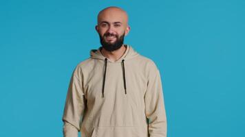 Middle eastern guy greeting people in front of camera, waving hello and smiling over blue background. Young person posing with confidence in studio, feeling confident and joyful. Camera 1. video