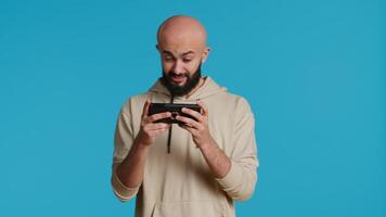 alegre musulmán hombre jugando móvil vídeo juegos en estudio, disfrutando en línea competencia utilizando teléfono inteligente aplicación Arábica jugador teniendo divertido con juego de rol desafío concurso. cámara 1. video