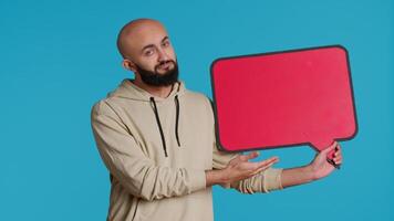 Arab person holding red speech bubble icon in studio, creating web advertisement for new product commercial. Middle eastern man shows isolated cardboard sign for ad. Camera 1. video