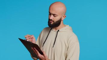 Middle eastern adult creating a checklist on clipboard papers, being focused on trying to plan tasks on an organizer in studio. Modern person looking at files and listing things to do. Camera 2. video