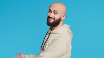 Smiling person advertising something on camera, pointing to left or right sides in studio. Middle eastern guy showing advertisement for marketing campaign, blue background. Camera 2. Handheld shot. video