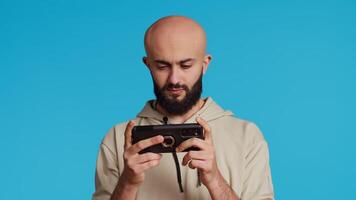 Middle eastern adult enjoying video games challenge in studio, playing online contest on smartphone with friends. Arab guy having fun with gaming shooter competition. Camera 2.