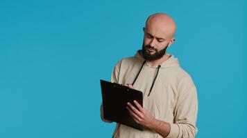 Middle eastern adult creating a checklist on clipboard papers, being focused on trying to plan tasks on an organizer in studio. Modern person looking at files and listing things to do. Camera 1. video