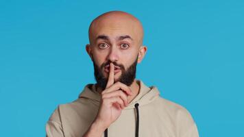 Middle eastern man doing hush silence symbol in studio, asking to keep a secret over blue background. Arab person creating mute gesture to be quiet in front of camera. Camera 2. video