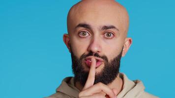 Middle eastern person showing mute gesture with finger over lips, posing over blue background. Arab guy doing hush symbol to keep silence, trying to share a secret whispering. Camera 2. video