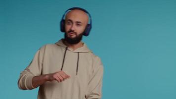 Muslim person listening to happy tunes on headphones, having fun with radio songs to feel relaxed and happy. Middle eastern guy dancing with headset over blue background. Camera 2. video