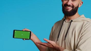 Middle eastern man presenting greenscreen on smartphone, showing chromakey display with isolated mockup layout in studio. Muslim adult creating promotion with blank copyspace screen. Camera 2. video