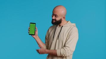 Muslim guy holding smartphone with greenscreen display, pointing at isolated chromakey template on mobile phone app. Young adult presenting blank copyspace mockup layout. Camera 1. video