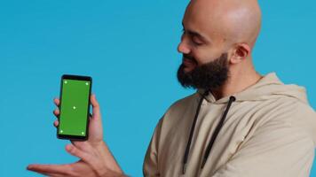 Muslim guy holding smartphone with greenscreen display, pointing at isolated chromakey template on mobile phone app. Young adult presenting blank copyspace mockup layout. Camera 2. video