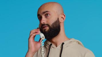 Muslim adult taking call on a landline phone in studio, using vintage office telephone with a cord. Arab guy standing over blue background and having a discussion on a remote call. Camera 2. video