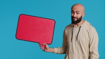 Arab person doing marketing ad with speech bubble, creating commercial with isolated empty billboard icon over blue background. Young adult holding cardboard sign for promo. Camera 1. video